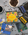 Overhead shot of a Coffee Tour box featuring women of Latin America; three coffee bags, with three info cards, a cup of coffee and beans scattered on a gray surface