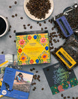 Overhead shot of a Coffee Tour box featuring women of Latin America; three coffee bags, with three info cards, a cup of coffee and beans scattered on a gray surface