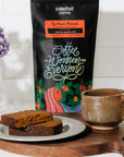 Coffee bag featuring women produced coffee from Rwanda, a mug with coffee, and desert bread on a white surface with flowers.