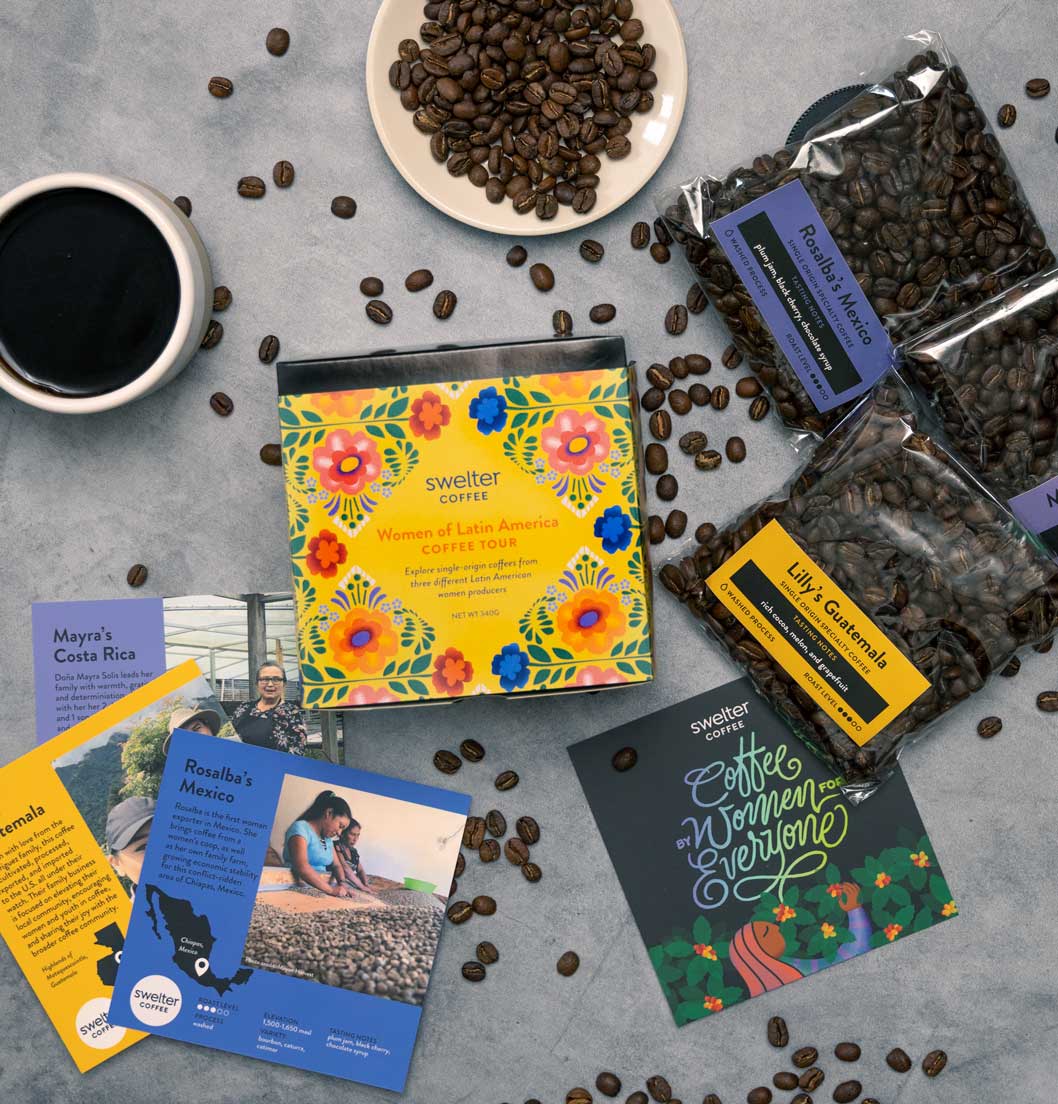 Overhead shot of a Coffee Tour box featuring women of Latin America; three coffee bags, with three info cards, a cup of coffee and beans scattered on a gray surface