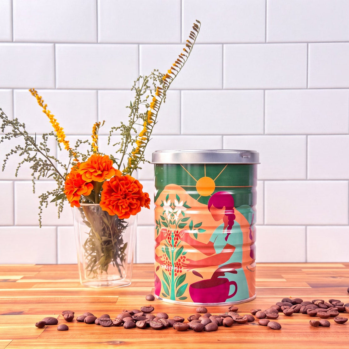 A coffee tin with an illustration of a woman picking coffee cherries, on a wood table with a vase of flowers next to it and coffee beans in the foreground.