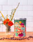 A coffee tin with an illustration of a woman picking coffee cherries, on a wood table with a vase of flowers next to it and coffee beans in the foreground.