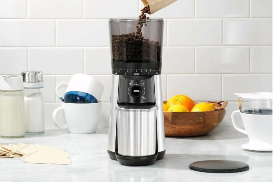 Coffee being poured into an Oxo Conical Grinder on a table in front of coffee mugs and a bowl of oranges. 