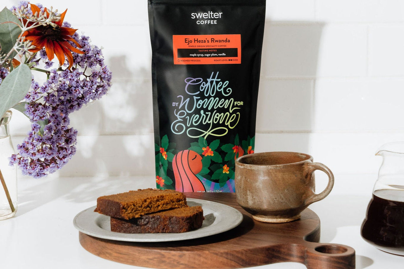 Coffee bag featuring women produced coffee from Rwanda, a mug with coffee, and desert bread on a white surface with flowers.