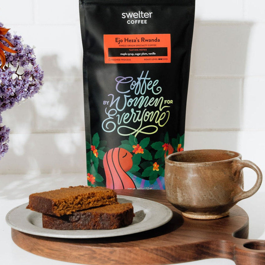 Coffee bag featuring women produced coffee from Rwanda, a mug with coffee, and desert bread on a white surface with flowers.