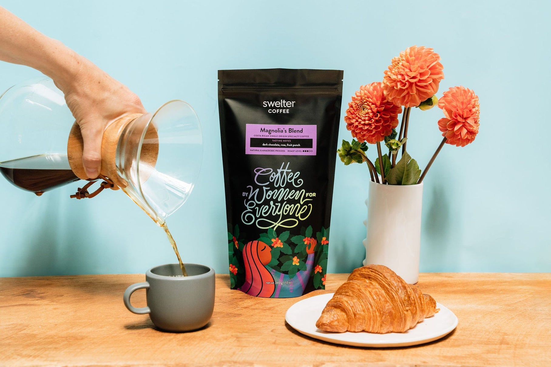 Coffee bag centered with a hand pouring coffee on the left, into a mug, and a vase with dalia flowers and a croissant on the right