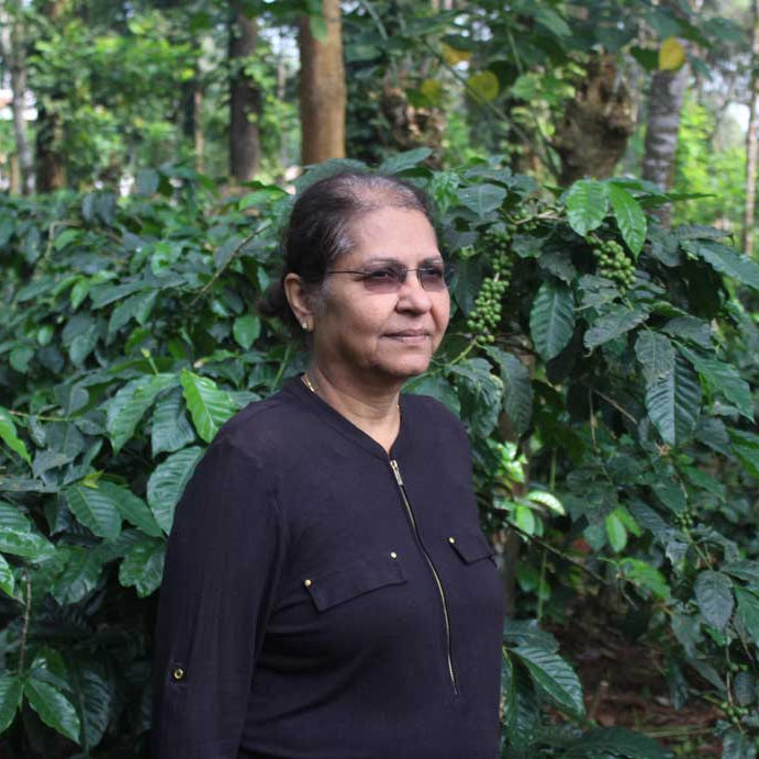 Woman coffee producer Poornima jairaj on her farm in southern India, standing in front of a few coffee trees with green cherries on them, under a larger tree canopy.