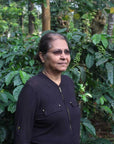 Woman coffee producer Poornima jairaj on her farm in southern India, standing in front of a few coffee trees with green cherries on them, under a larger tree canopy.